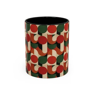 May include: A ceramic coffee mug with a black interior and a repeating geometric pattern. The pattern features red circles, green half-circles, and tan and brown curved shapes. The mug is cylindrical and sits on a white surface.