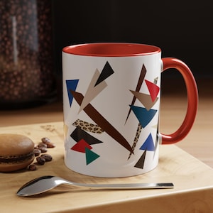 May include: White ceramic mug with a red interior and handle. The mug features a geometric design with triangles in various colors, including brown, blue, red, and green. A spoon and a macaron are on a wooden surface.