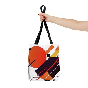 May include: A white tote bag with black straps and a geometric design featuring orange, red, yellow, and black shapes.