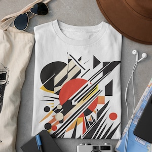 May include: White t-shirt with a geometric design featuring black, white, red, and yellow shapes. The design includes a large red circle and several smaller circles. The t-shirt is laid flat on a gray surface with other travel accessories.