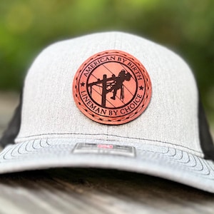 May include: A gray and black trucker hat with a circular leather patch. The patch features a lineman silhouette on a utility pole, with the text "AMERICAN BY BIRTH" and "LINEMAN BY CHOICE" in a circular design. The hat has a curved brim.