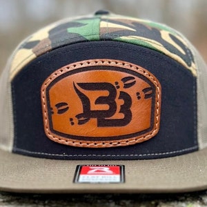May include: A camouflage, black, and brown baseball cap with a brown leather patch featuring a stylized "BB" logo. The cap has a mesh back and a flat brim.