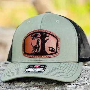 May include: A green and black trucker hat with a brown leather patch featuring a dog and a cat in a tree. The patch has the text "B" on it. The hat has a black mesh back and a green front with the text "Richardson. Est. 1970 Original Trucker 112 Adjustable" on the front.
