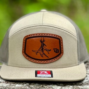 May include: A tan and gray flat bill baseball cap with a brown leather patch featuring a deer antler design and the letter "B". The patch has stitching around the edges. The cap has a mesh back and a flat bill.