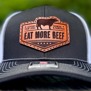May include: Black and white trucker hat with a leather patch. The patch features a brown bull silhouette and the text "EAT MORE BEEF" with "SUPPORT YOUR LOCAL FARMERS & RANCHERS". The hat has a curved brim and a mesh back.