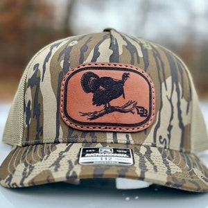 May include: A camouflage trucker hat with a brown leather patch featuring a black silhouette of a turkey. The patch has the text "EST. 1970 RICHARDSON 112" on the bottom.