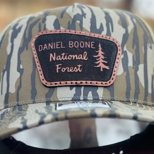 May include: A camouflage trucker hat with a brown leather patch that says "Daniel Boone National Forest" in a vintage font with a pine tree graphic.