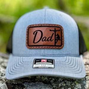 May include: A gray and black trucker hat with a brown leather patch that says "Dad" with a silhouette of a lineman working on a power line.