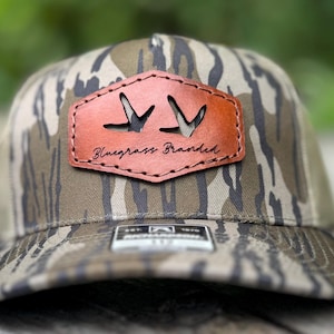 May include: A camouflage-patterned baseball cap with a brown leather patch. The patch features two cut-out bird silhouettes and the text "Bluegrass Branded" in a script font. The cap has a curved brim.