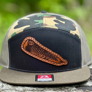May include: A black and green camouflage flat bill baseball cap with a brown leather patch on the front. The patch has a detailed design of a fish.