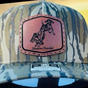 May include: A camouflage baseball cap with a brown leather patch featuring a black and white illustration of a dog chasing a rabbit. The patch has the text "Bluegrass Branded" on it.