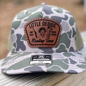 May include: A camouflage trucker hat with a brown leather patch that says "Little Debbie Hunting Team" with a cartoon girl holding a rifle and a snack. The hat is adjustable and has a Richardson logo.
