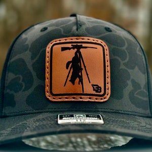 May include: Black camouflage trucker hat with a brown leather patch featuring a silhouette of a person in a hunting blind with a rifle.