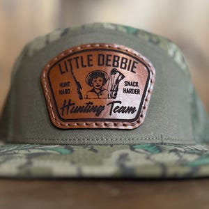 May include: A green camouflage baseball cap with a brown leather patch that says "Little Debbie Hunting Team" with a cartoon girl holding a rifle and bow and arrow. The patch also says "Hunt Hard" and "Snack Harder".