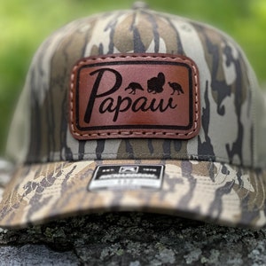 May include: Camouflage baseball cap with a brown leather patch. The patch features the word "Papaw" and silhouettes of turkeys. The cap has a curved brim and a snapback closure.