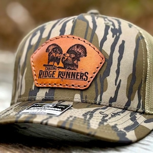 May include: A camouflage baseball cap with a brown leather patch featuring two turkey illustrations and the text "Chasing Ridge Runners." The cap has a mesh back and a curved brim. The hat is designed with a hunting theme.