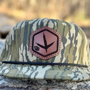 May include: A camouflage baseball cap with a brown leather patch featuring a black bird's footprint and the letter "B".