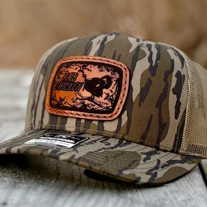 May include: A camouflage trucker hat with a brown leather patch featuring a turkey illustration and the text "Limb Chicken." The hat has a curved brim and a mesh back.