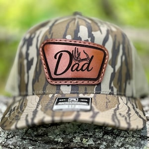 May include: A camouflage trucker hat with a brown leather patch that says "Dad" with a deer antler design.