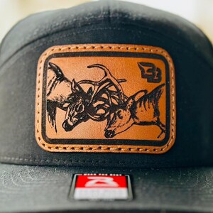 May include: Black trucker hat with a brown leather patch featuring a black and white illustration of two deer with antlers locked in a fight. The patch has the letter "B" in a circle on the right side. The hat has a black mesh back.