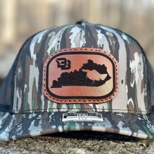 May include: A camouflage trucker hat with a brown leather patch featuring the outline of the state of Kentucky and the letters "B3".