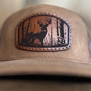 May include: A brown and black trucker hat with a brown leather patch featuring a silhouette of a deer in a forest setting.