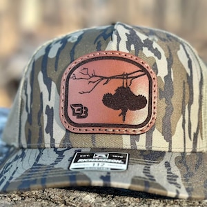 May include: A camouflage trucker hat with a brown leather patch featuring a silhouette of a bird perched on a branch. The patch has the letter "B" in a circle on the left side.