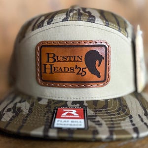 May include: A camouflage patterned baseball cap with a brown leather patch that says "Bustin Heads'25" and a black silhouette of a fish. The cap has a flat bill and a "Remarkable" logo on the side.