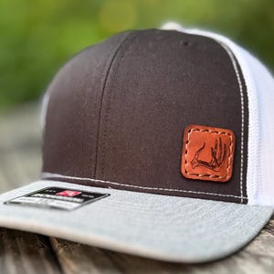 May include: A brown and white trucker hat with a gray brim. The hat features a leather patch with a deer antler design. The hat is sitting on a wooden surface.