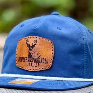May include: A blue baseball cap with a flat brim and a white rope detail. The cap features a brown leather patch with a deer graphic and the words "BLUEGRASS BRANDED".