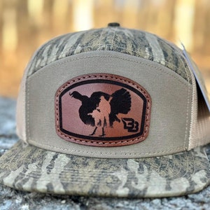 May include: A brown and tan camouflage trucker hat with a brown leather patch featuring a silhouette of a turkey and a hunter. The patch has the text "B3" on it.