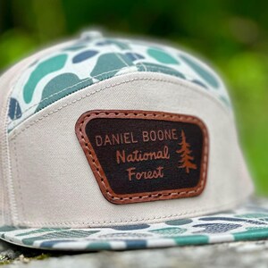 May include: A beige and green camouflage patterned baseball cap with a brown leather patch that says "Daniel Boone National Forest".