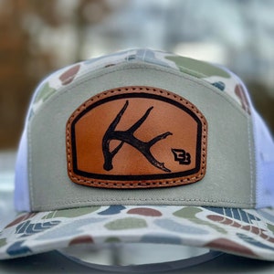 May include: A gray and white camouflage trucker hat with a brown leather patch featuring a stylized antler design and the letter "B".