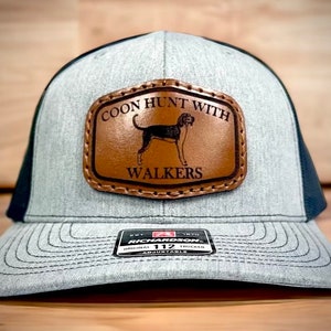 May include: A gray and black trucker hat with a brown leather patch that says "Coon Hunt With Walkers" and features a black and brown hound dog. The hat is adjustable and has a black mesh back.
