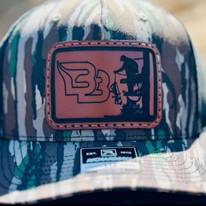 May include: A camouflage trucker hat with a brown leather patch featuring a silhouette of a person in a tree stand and the text "B3". The hat is made by Richardson and has the text "Est. 1970" on the back.
