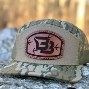 May include: A camouflage patterned baseball cap with a brown leather patch featuring a black stylized letter "B" with arrows pointing towards it.