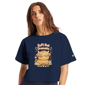 May include: A navy blue short-sleeved t-shirt featuring a graphic of a fluffy highland cow with a pink bow. The text above the cow reads "Soft But Dramatic" in a light-colored font, surrounded by small star and flower illustrations.