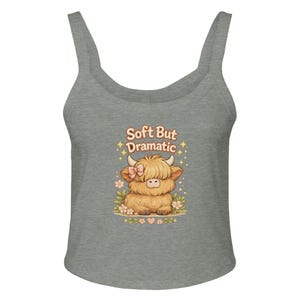 May include: A heather gray tank top featuring a graphic of a fluffy tan highland cow with a pink bow. The cow is surrounded by small flowers and stars. Above the cow, the text reads "Soft But Dramatic" in a rounded, orange-toned font.