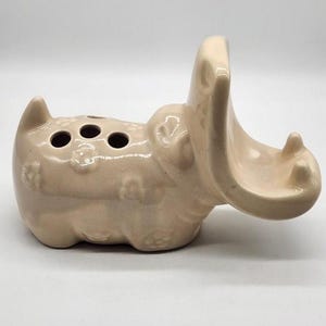 May include: A light beige ceramic hippopotamus figurine with a glossy finish. The hippo has its mouth wide open and features three circular holes on its back, along with embossed flower patterns across its body and a small pointed tail.