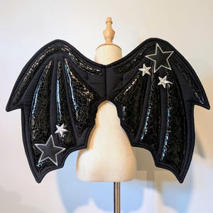 May include: Black bat-style wings featuring a glossy, cracked-texture finish on the panels. The wings are decorated with white and black star-shaped appliqués with visible stitching along the edges, mounted on a beige mannequin torso.