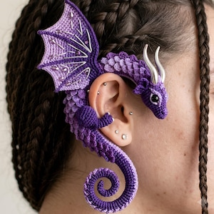 May include: A purple crocheted dragon ear cuff featuring a textured scale pattern, a large wing with silver-toned embroidery, and two smooth silver-toned horns. The dragon body curves around the ear, ending in a spiral tail at the bottom.