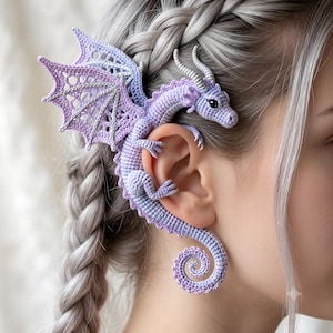 May include: A lavender and light purple crocheted dragon ear cuff featuring intricate wing detailing with silver thread accents, a curled tail, and small horns. The dragon is designed to wrap around the outer edge of the ear structure.