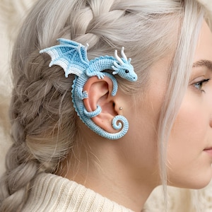 May include: A light blue and white crocheted dragon ear cuff that wraps around the outer ear. The dragon features detailed wings, a curved tail, and small white horns. The texture is made of fine yarn stitches, creating a soft, intricate appearance.