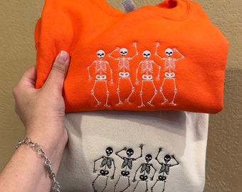 May include: Two sweatshirts, one orange and one beige, each embroidered with a line of dancing skeletons. The orange sweatshirt has white skeleton embroidery, while the beige one has black skeleton embroidery. The sweatshirts are folded and stacked.