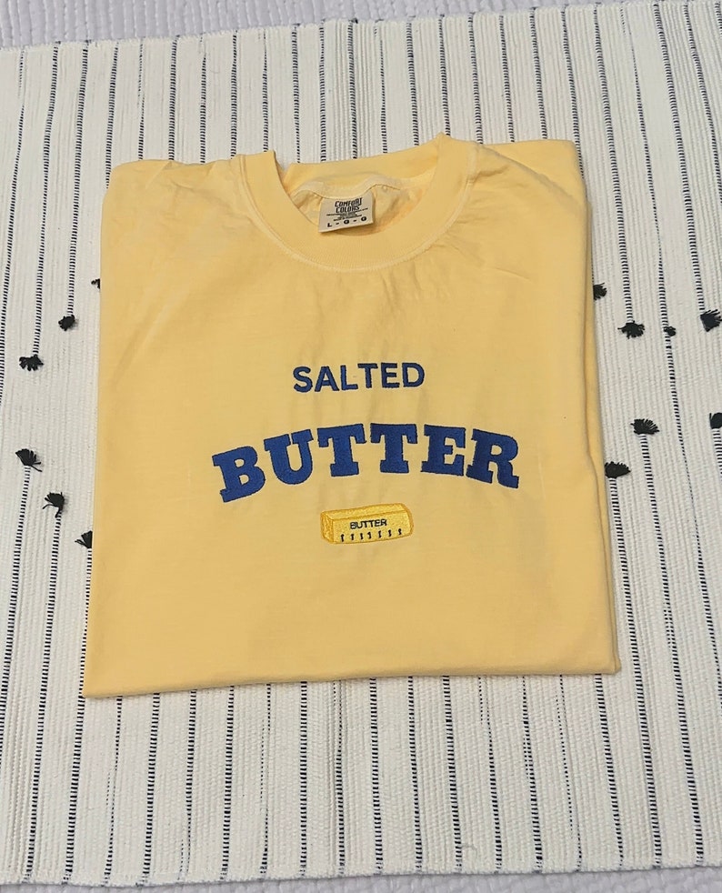 Pu&ograve; includere: Una maglietta gialla con le parole "SALTED BUTTER" in lettere blu. Sotto la parola "BUTTER" c'&egrave; una piccola grafica di un panetto di burro. La maglietta &egrave; piegata su una superficie a righe bianche e nere.