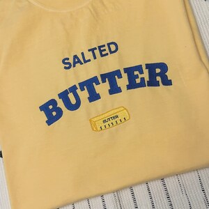 Pu&ograve; includere: Una felpa giallo chiaro con le parole "SALTED BUTTER" in blu, a forma di arco. Sotto il testo c'&egrave; una piccola grafica ricamata di un panetto di burro. La felpa &egrave; piegata su una superficie a righe.