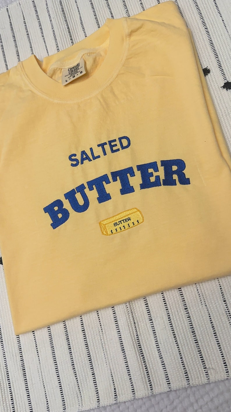 Pu&ograve; includere: Una felpa giallo chiaro con le parole "SALTED BUTTER" in blu, a forma di arco. Sotto il testo c'&egrave; una piccola grafica ricamata di un panetto di burro. La felpa &egrave; piegata su una superficie a righe.