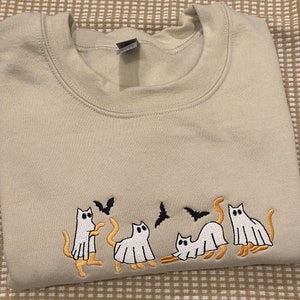 May include: Beige sweatshirt with an embroidered Halloween design. The design features four white ghost cats with orange tails and legs, and three black bats. The sweatshirt is folded neatly.