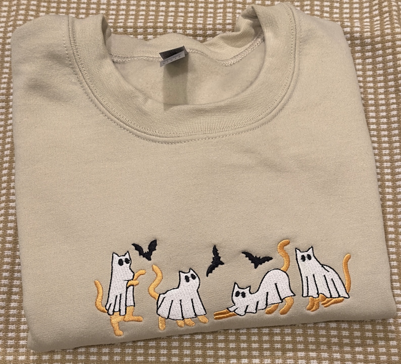 May include: Beige sweatshirt with an embroidered Halloween design. The design features four white ghost cats with orange tails and legs, and three black bats. The sweatshirt is folded neatly.