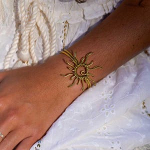 Könnte beinhalten: Ein goldfarbenes Armband mit Sonnenstrahlen. Das Armband hat eine spiralförmige Mitte mit strahlenförmigen Linien, die ein sonnenähnliches Design bilden. Der Schmuck wird am Handgelenk getragen, vor einem weißen Stoffhintergrund.